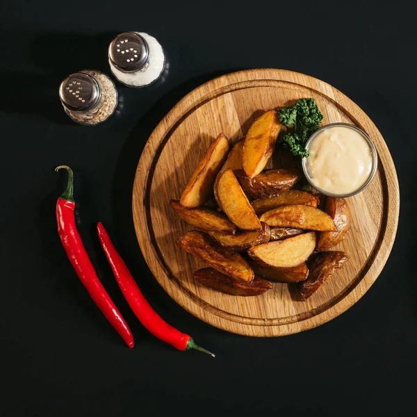 Leckere Ofenkartoffeln mit Sauce auf Holzbrett, Gewürze und Chilischoten auf schwarzem — Stockfoto