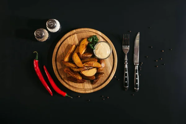 Tasty roasted potatoes with sauce on wooden board, spices, chili peppers and fork with knife on black — Stock Photo