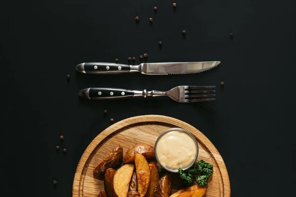 Top view of tasty roasted potatoes with sauce on wooden board and fork with knife on black — Stock Photo