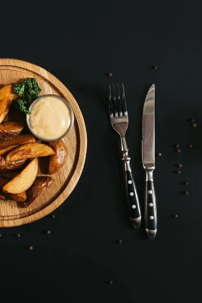 Top view of baked potatoes with sauce on wooden board and fork with knife on black — Stock Photo