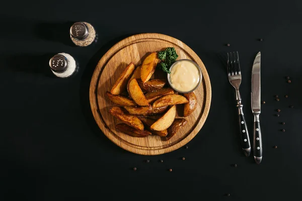 Top view of baked potatoes with sauce on wooden board, fork with knife and spices on black — Stock Photo