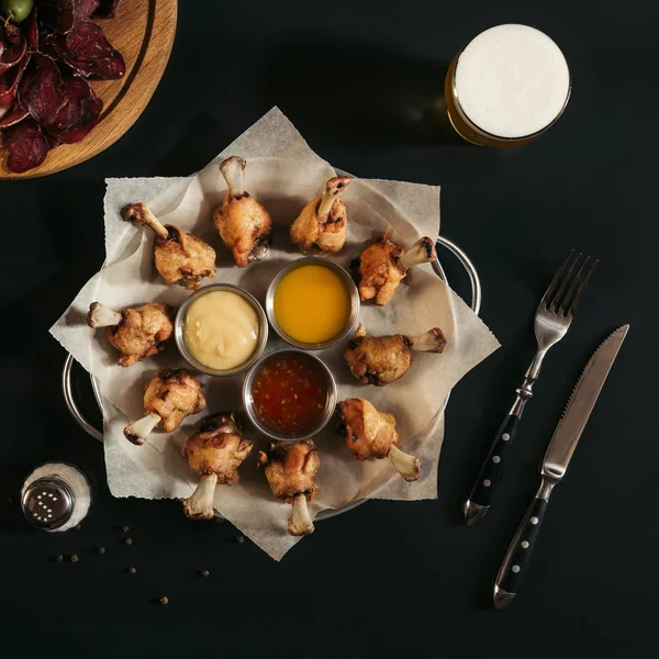 Vista superior de deliciosas alitas de pollo asado con varias salsas y vaso de cerveza en negro - foto de stock