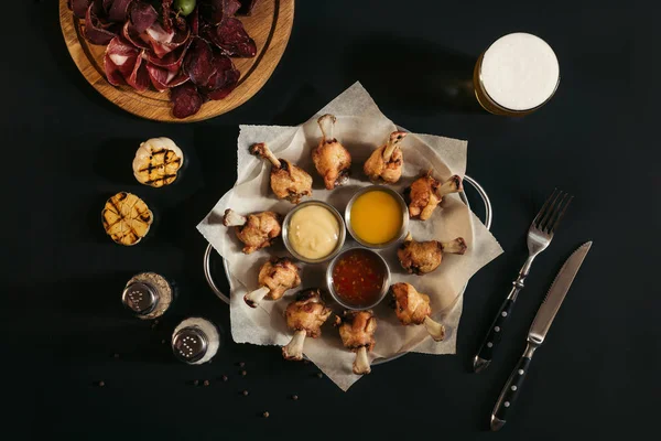 Жареная курица с соусами, специями, стаканом пива и разнообразным ремесленным мясом на черном — стоковое фото