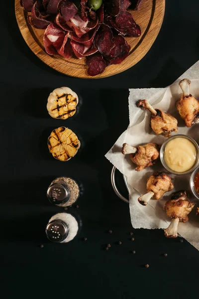 Top view of fried tasty chicken wings with sauces, spices, grilled garlic and gourmet assorted craft meat on black — Stock Photo