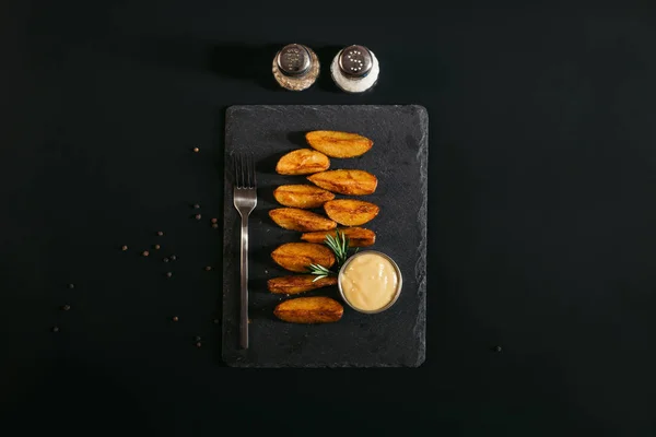 Вид на вкусную выпечку картофеля с соусом и вилкой на доске и специи на черном — стоковое фото