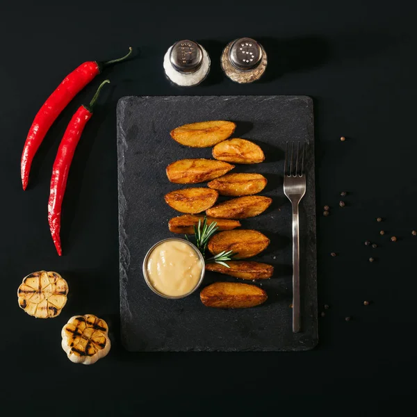 Leckere Ofenkartoffeln mit Sauce und Gabel auf Schiefertafel und Gewürzen mit Knoblauch und Chilischoten auf Schwarz — Stockfoto