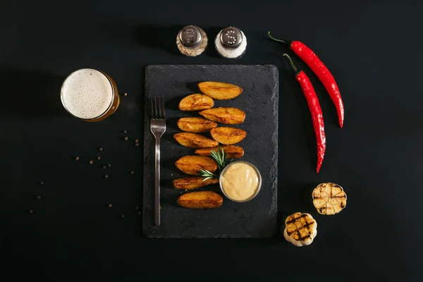 Vue de dessus des pommes de terre cuites au four savoureuses avec sauce et fourchette sur ardoise et verre de bière sur noir — Photo de stock