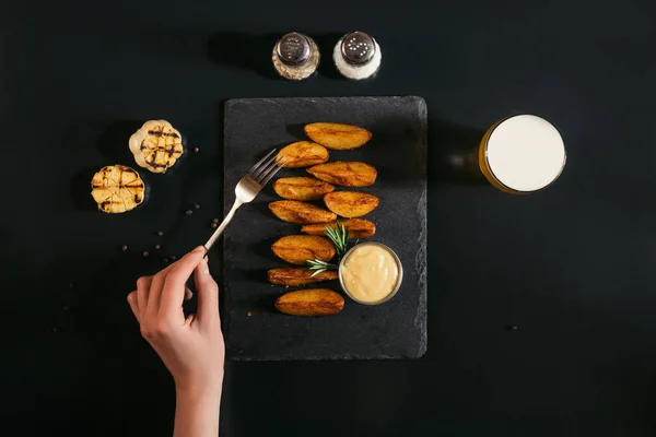 Partial top view of person holding fork and eating tasty baked potatoes with sauce on black — Stock Photo