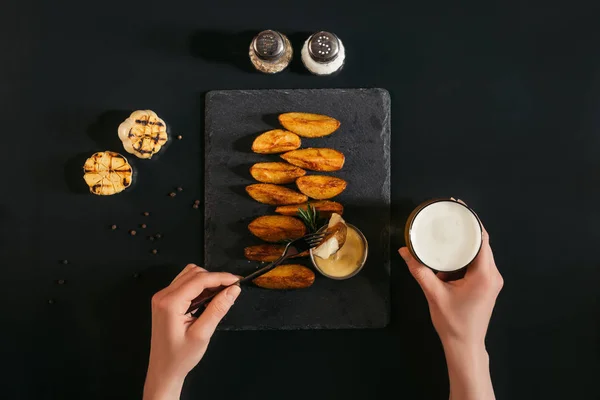 Cropped shot of person drinking beer and eating baked potatoes with sauce on black — Stock Photo