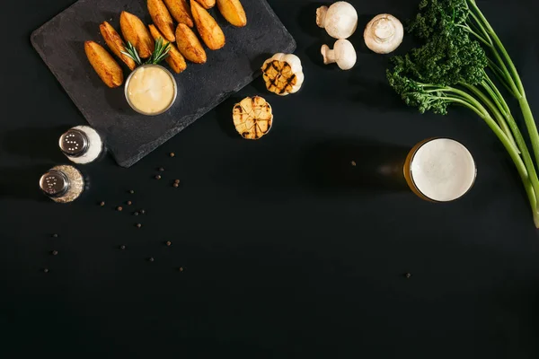 Vue du dessus de délicieuses pommes de terre cuites au four avec sauce, épices et verre de bière sur noir — Photo de stock