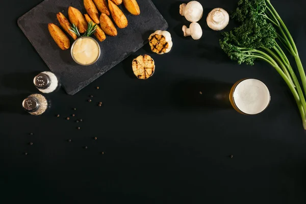 Vista superior de deliciosas papas al horno con salsa, especias y vaso de cerveza en negro - foto de stock