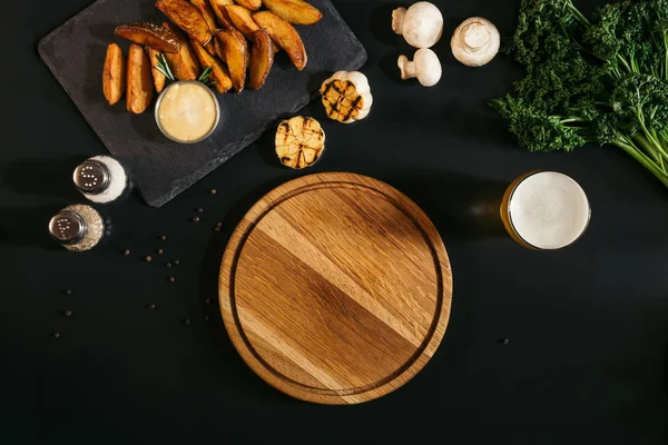 Leere Holzplatte, Glas Bier und Bratkartoffeln mit Sauce und Gewürzen auf Schwarz — Stockfoto