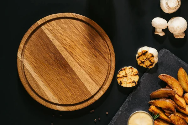 Vue du dessus de la planche de bois vide, ail grillé, champignons et pommes de terre cuites au four avec sauce sur noir — Photo de stock