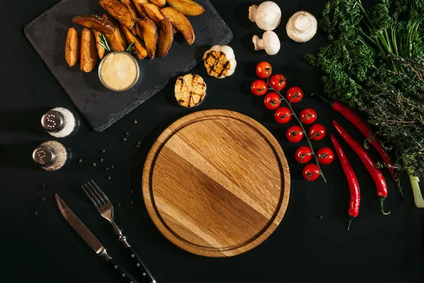 Top view of empty wooden board, vegetables and baked potatoes on black — Stock Photo