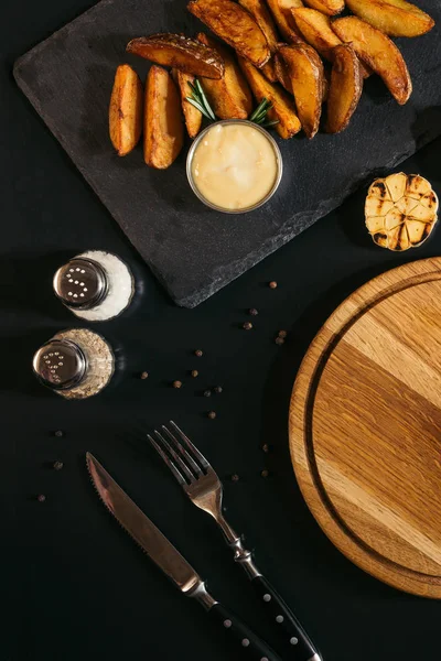 Вид сверху на вкусную выпечку картофеля с соусом, специями, жареным чесноком и деревянной доской с вилкой и ножом на черном — стоковое фото