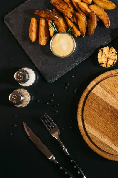 Vista superior de sabrosas papas al horno con salsa, especias, ajo a la parrilla y tabla de madera con tenedor y cuchillo en negro - foto de stock