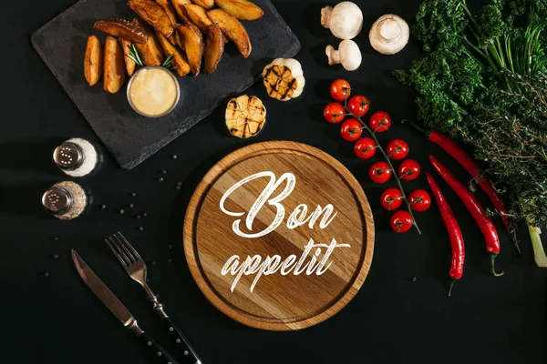 Wooden board with inscription bon appetit, baked potatoes and vegetables on black — Stock Photo