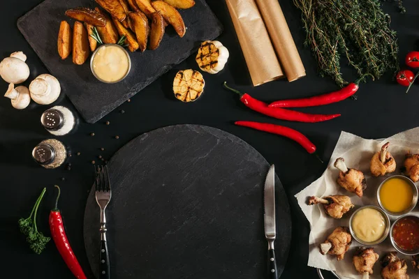 Vista superior de pizarra vacía con tenedor y cuchillo, patatas asadas y verduras en negro - foto de stock