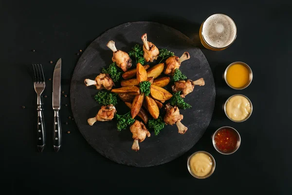 Patatas horneadas gourmet con pollo, vaso de cerveza, tenedor con cuchillo y varias salsas en negro - foto de stock