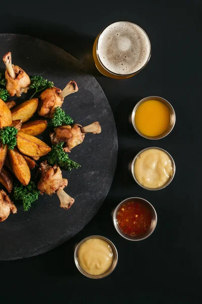 Vista superior de papas horneadas gourmet con pollo, vaso de cerveza y varias salsas en negro - foto de stock