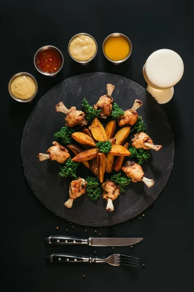 Délicieuses pommes de terre au four avec poulet, diverses sauces, fourchette avec couteau et verre de bière sur noir — Photo de stock