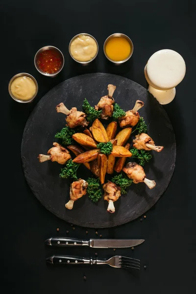 Deliciosas batatas assadas com frango, vários molhos, garfo com faca e copo de cerveja em preto — Fotografia de Stock