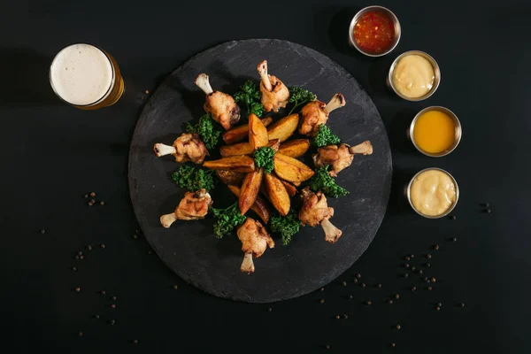 Blick von oben auf leckere Ofenkartoffeln mit Huhn, verschiedenen Soßen und einem Glas Bier auf schwarz — Stockfoto