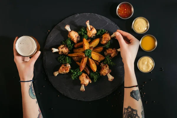 Cropped shot of person eating roasted chicken with potatoes and drinking beer on black — Stock Photo