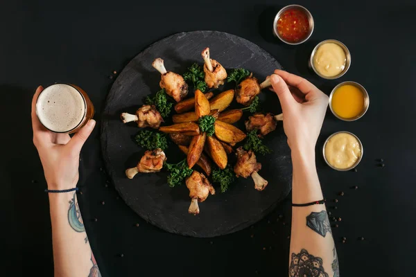 Tiro cortado de pessoa comendo frango assado com batatas e beber cerveja em preto — Fotografia de Stock