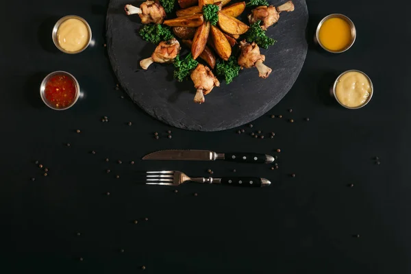 Вид сверху на вкусную выпечку картофеля с жареной курицей, различные соусы и вилку с ножом на черном — стоковое фото
