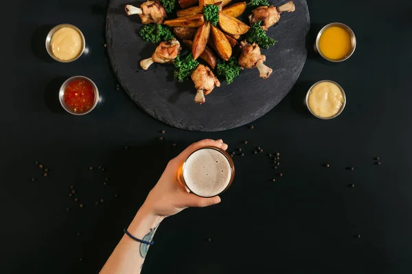 Plan recadré de la personne tenant un verre de bière tout en mangeant de délicieuses pommes de terre rôties avec du poulet et des sauces sur noir — Photo de stock