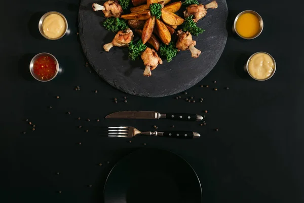 Top view of tasty baked potatoes with fried chicken wings, sauces and empty plate on black — Stock Photo