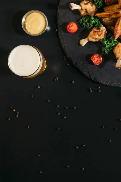 Вид сверху на пиво, соус и вкусную выпечку картофеля с жареными куриными крылышками на черном — стоковое фото