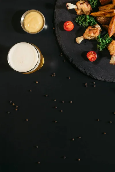 Vue du dessus du verre de bière, sauce et savoureuses pommes de terre cuites au four avec des ailes de poulet frit sur noir — Photo de stock