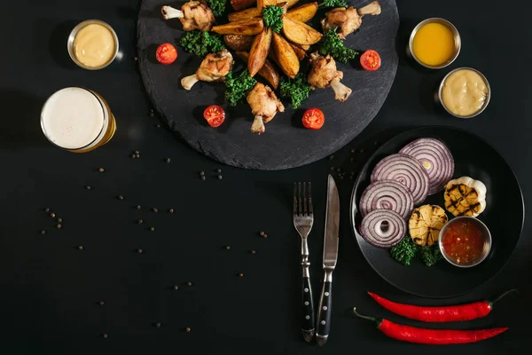 Blick von oben auf schmackhafte Bratkartoffeln mit Chicken Wings, Soßen, Gemüse und einem Glas Bier auf Schwarz — Stockfoto