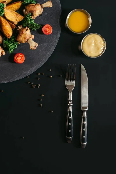 Вид зверху виделка з ножем і смачна смажена картопля з курячими крильцями і соусами на чорному — стокове фото