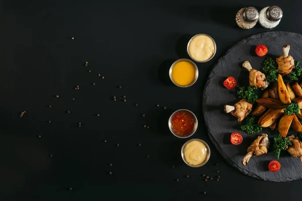 Verschiedene Soßen, Gewürze und leckere Ofenkartoffeln mit gebratenen Hühnerflügeln auf Schiefertafel auf schwarz — Stockfoto