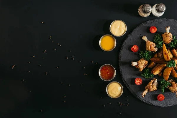 Varie salse, spezie e deliziose patate al forno con ali di pollo arrosto su tavola di ardesia su nero — Foto stock