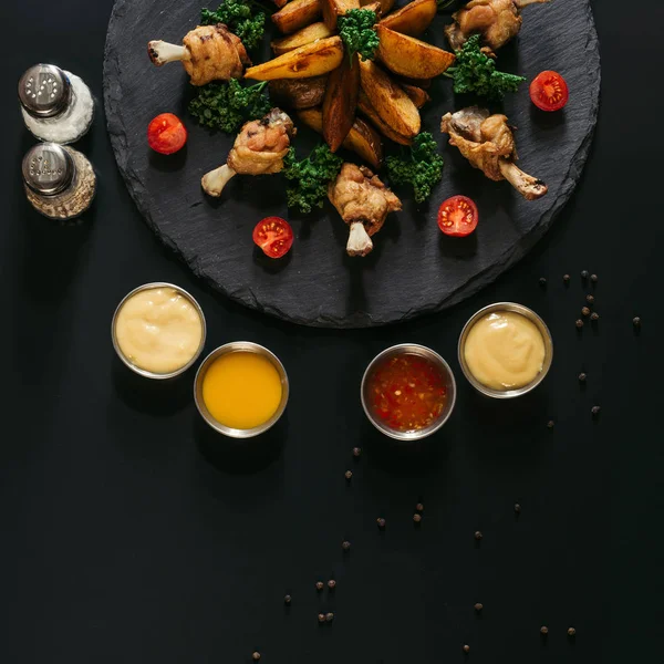 Vue de dessus de diverses sauces, épices et délicieuses pommes de terre cuites au four avec des ailes de poulet rôties sur noir — Photo de stock