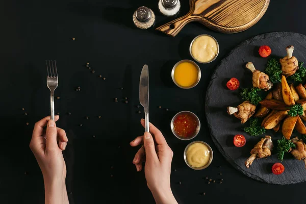 Vista superior parcial de la persona sosteniendo tenedor y cuchillo y papas horneadas sabrosas con pollo asado y salsas en negro - foto de stock