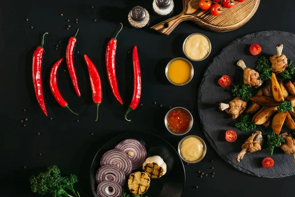 Vista superior de pimentas vermelhas, molhos, legumes e batatas assadas saborosas com frango assado em preto — Fotografia de Stock