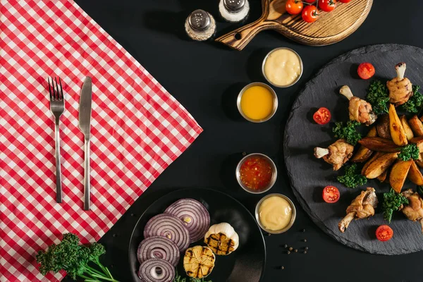 Vue du dessus de la serviette à carreaux avec fourchette et couteau, légumes, sauces et délicieuses pommes de terre rôties avec poulet sur noir — Photo de stock