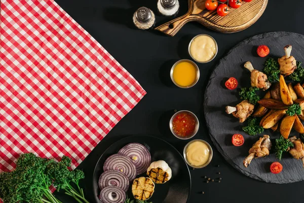 Vista superior de servilleta a cuadros, verduras, salsas y deliciosas papas asadas con pollo sobre negro - foto de stock