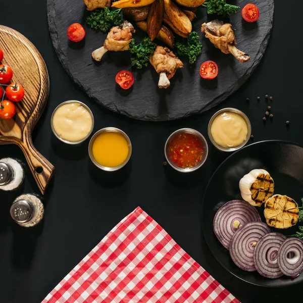 Vue de dessus de diverses sauces, de délicieuses ailes de poulet et de pommes de terre cuites au four sur noir — Photo de stock