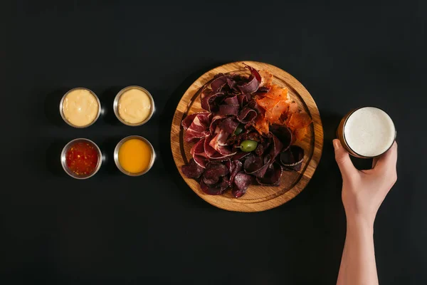 Cropped shot of person holding glass of beer and delicious sliced assorted meat on wooden board with various sauces on black — Stock Photo