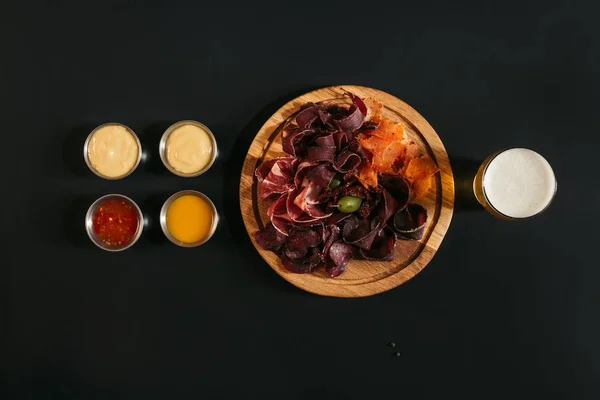 Vista superior de deliciosa carne rebanada surtida en tablero de madera con vaso de cerveza y salsas en negro - foto de stock