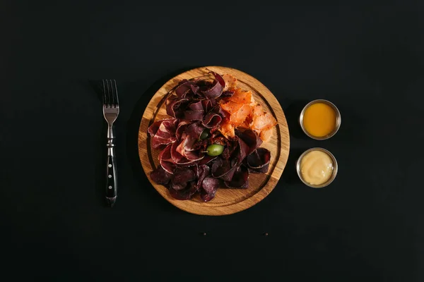 Top view of delicious sliced assorted meat on wooden board with fork and sauces on black — Stock Photo