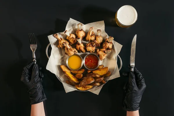 Plan recadré de la personne dans des gants tenant fourchette et couteau tout en mangeant des pommes de terre cuites au four avec des sauces et du poulet rôti sur noir — Photo de stock