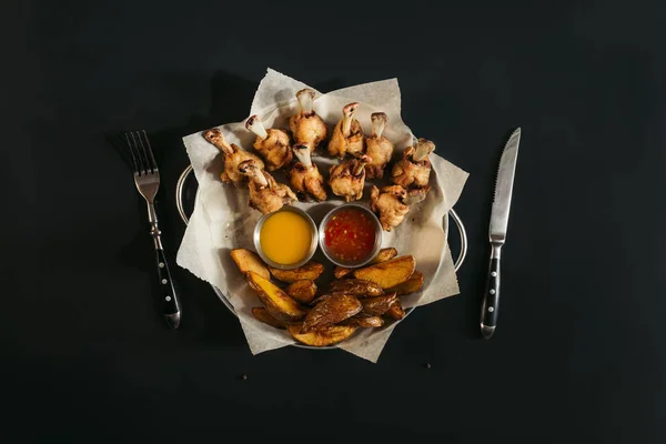 Vue du dessus de délicieuses pommes de terre cuites au four avec des sauces et des ailes de poulet rôties sur noir — Photo de stock
