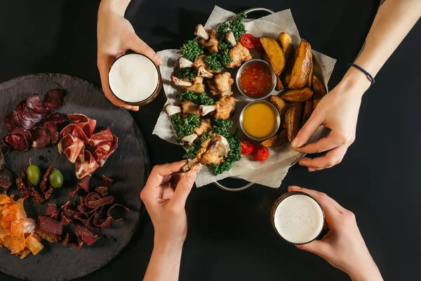 Plan recadré de personnes buvant de la bière et mangeant de délicieuses pommes de terre au four avec du poulet rôti sur noir — Photo de stock